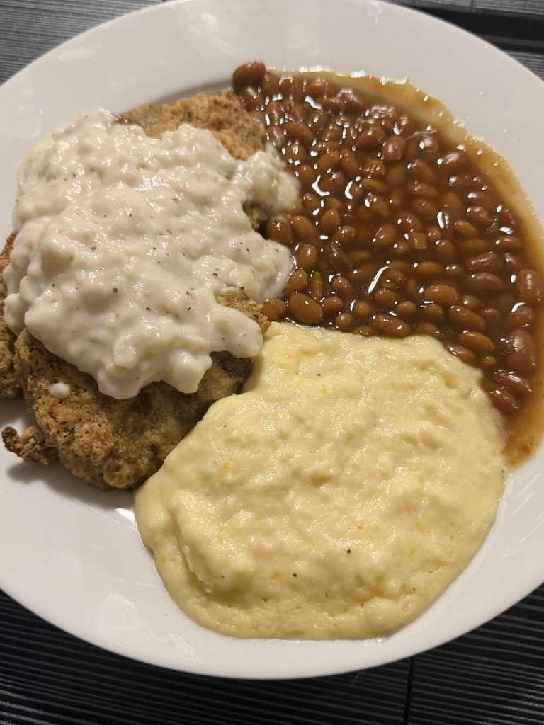 Country fried steak with creamy gravy, baked beans, protein mashed potatoes, and cornbread on a plate.