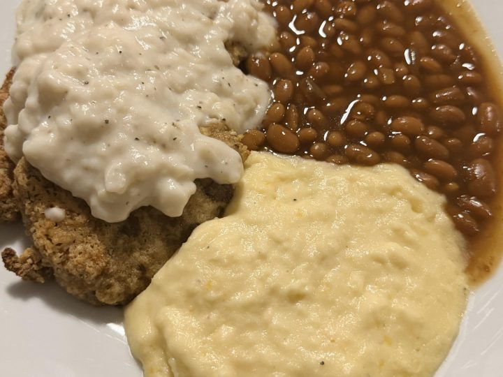 Country fried steak with creamy gravy, baked beans, protein mashed potatoes, and cornbread on a plate.